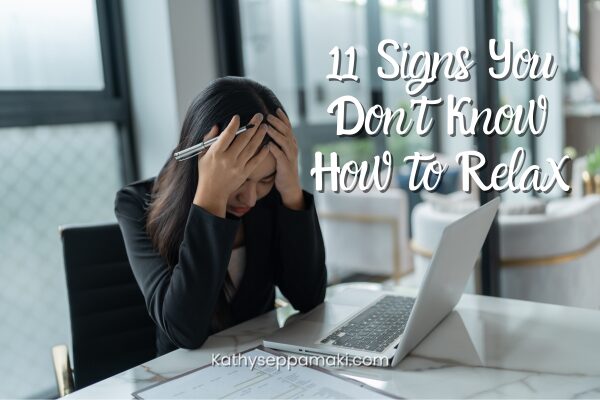 11 Signs You Don’t Know How to Relax blog post title with picture of a woman sitting at a table in front of her laptop looking stressed and holding her head in her hands.
