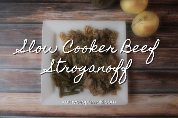 Slow Cooker Beef Stroganoff blog post title with photo of a plate of beef stroganoff over pasta and green beans. They are on a white square plate sitting on a wooden table top.